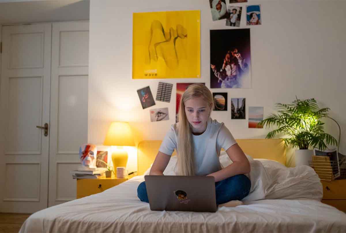دختری که با لپ تاپ باز روی تخت اتاقش نشسته