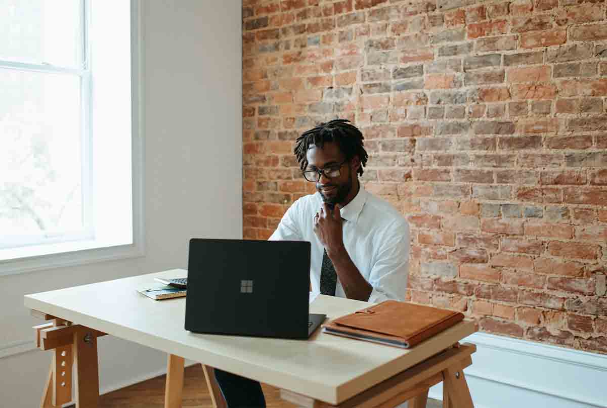 اقای سیاه پوست در حال کار با لپ تاپ پشت میز کار و دیوار اجری