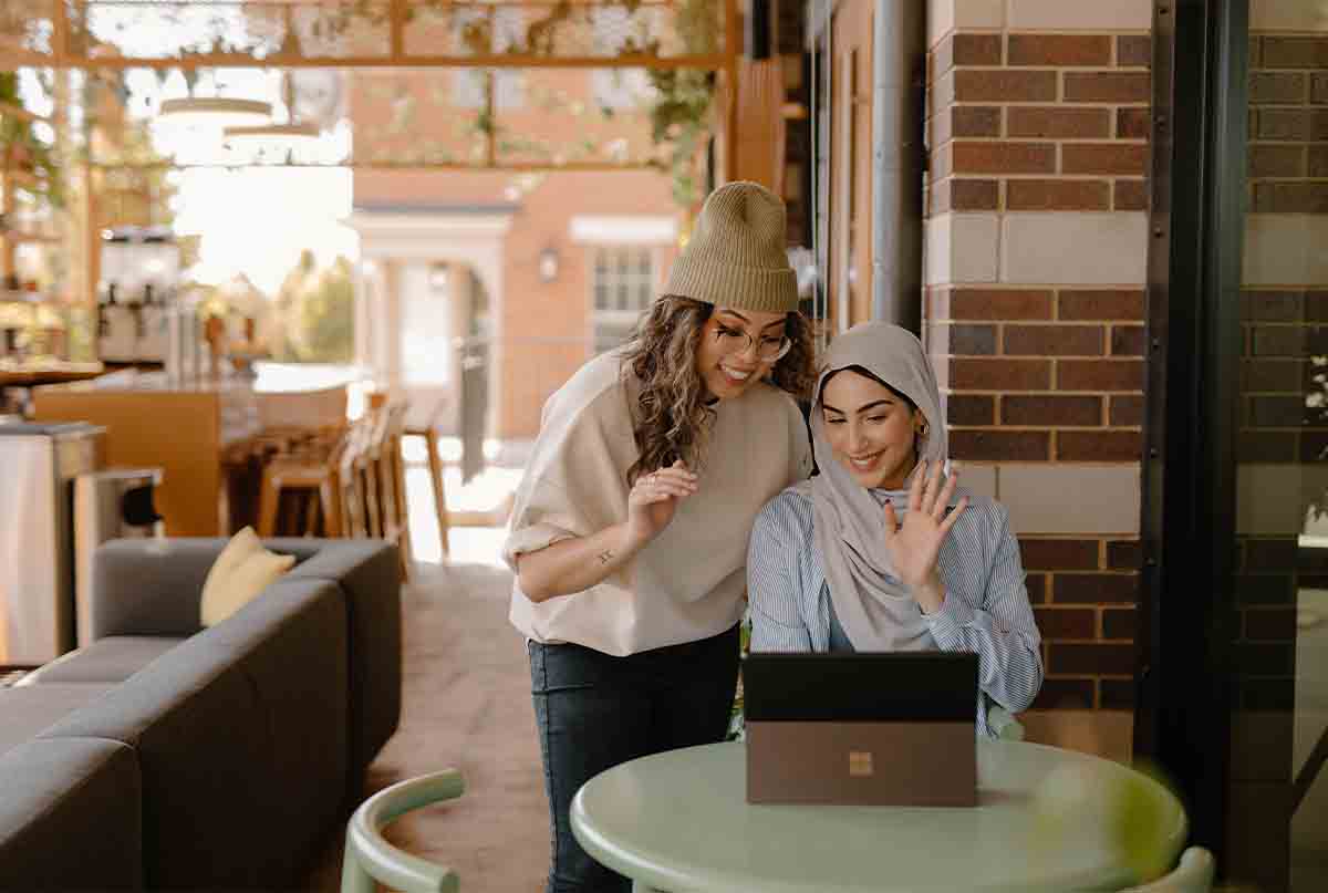 یک خانم با شال نشسته و یک خانم با کلاه ایستاده و به لپ تاپ نگاه میکنند