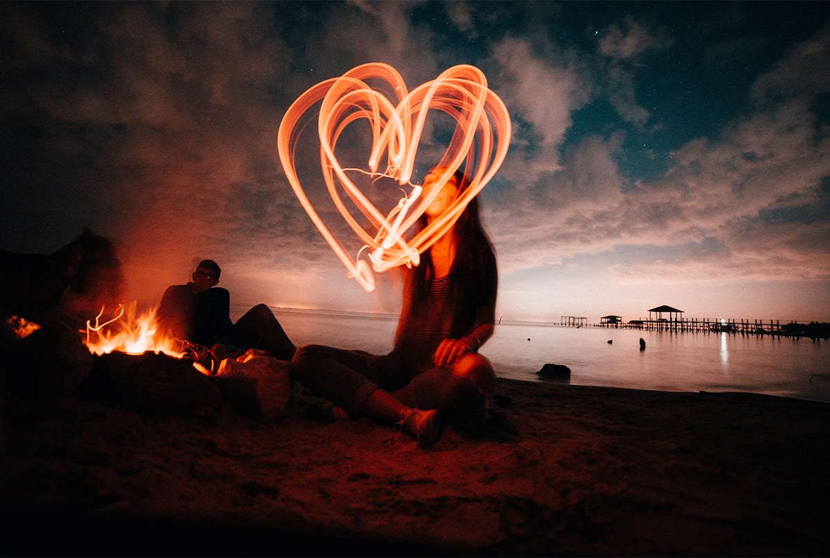 دختری که کنار ساحل نشسته و با نور تو اسمون قلب میکشه