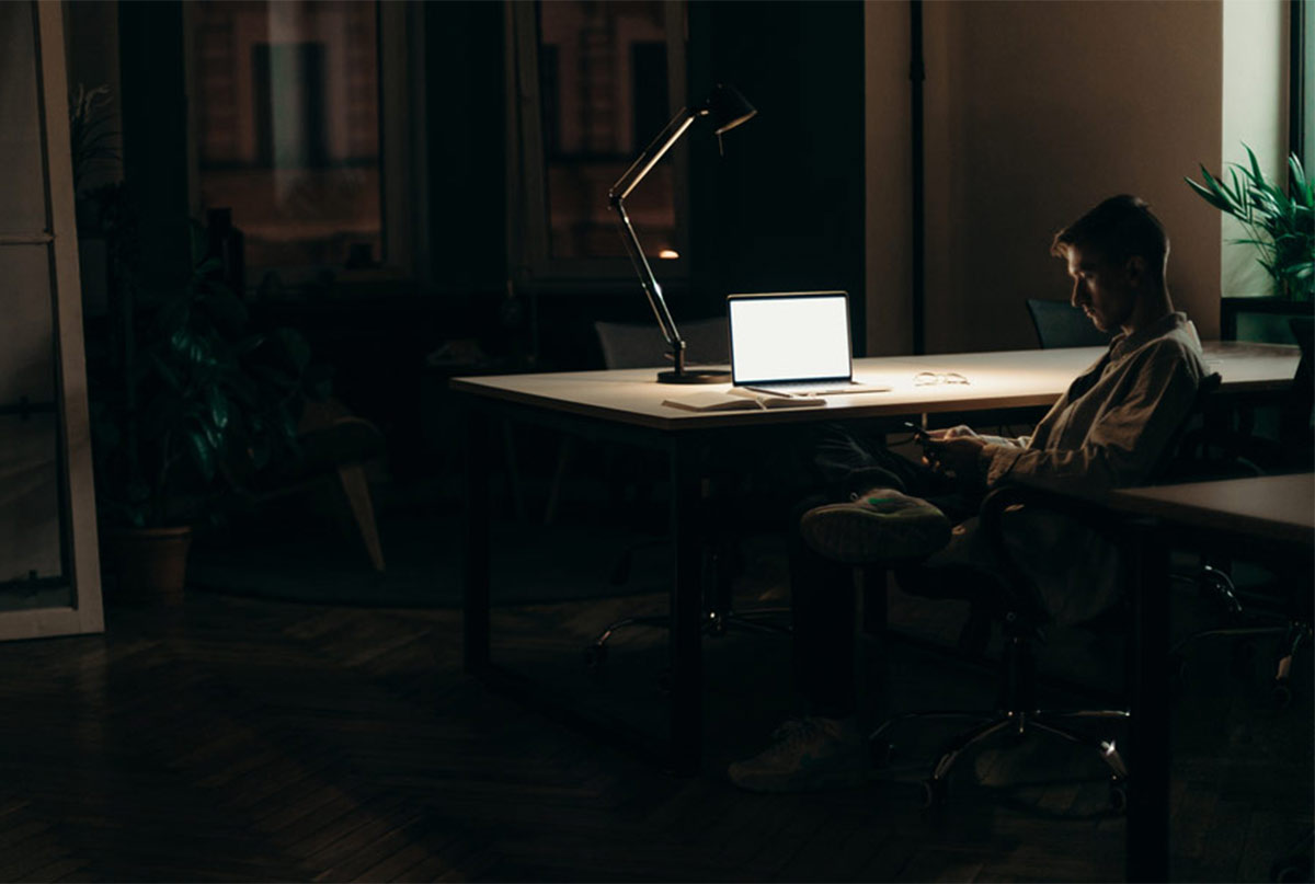 اقایی که پشست میز در اتاقی تاریک نشسته و لپ تاپش روشنه و خودش با گوشی کار میکنه