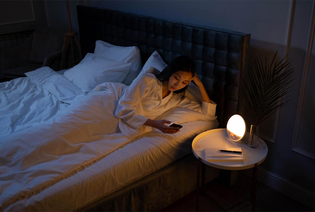 خانمی که تو تخت دراز کشیده و شبه و داره با گوشیش کار میکنه و چراغ خواب کنارش هم روشنه