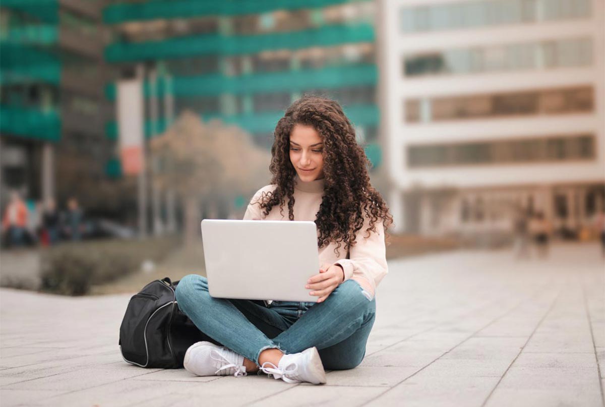 دختری با موهای فر که شلوار لی پوشیده و لپ تاپ دستشه و وسط محوطه ای نشسته و کولش هم کنارشه